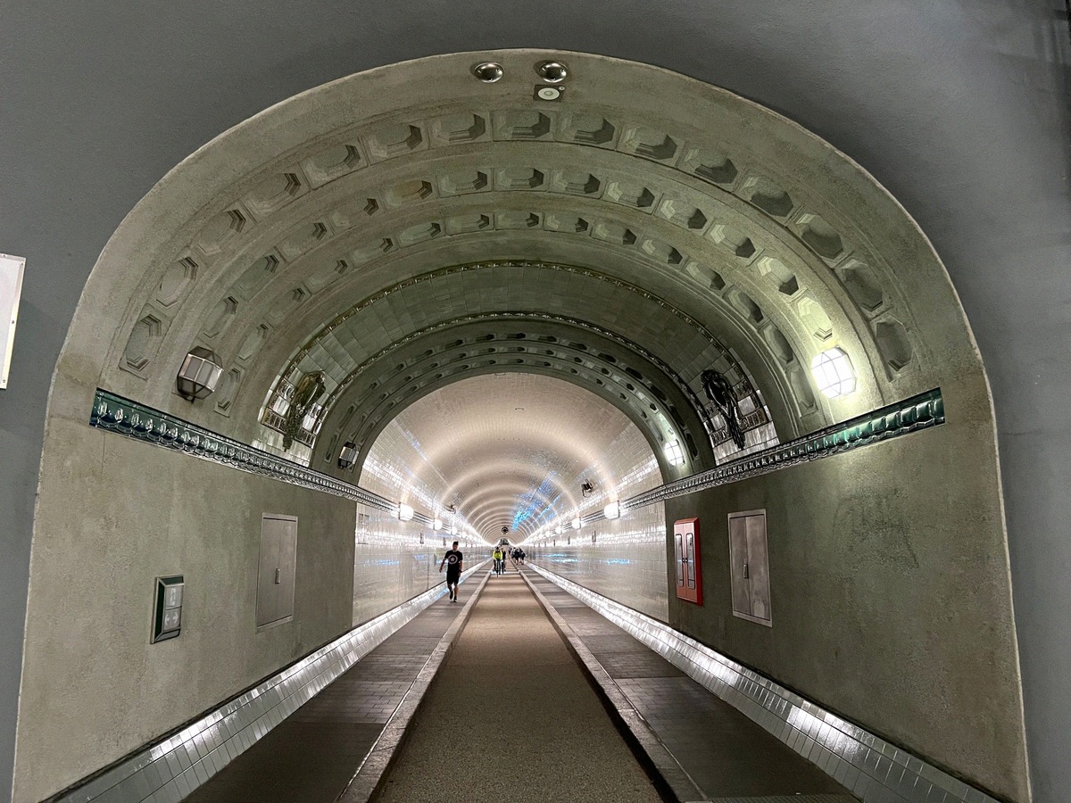 Symmetrische Innenaufnahme des Alten Elbtunnels: gewölbter Betongang mit Jugendstildekor, blaues Licht am Ende, einzelner Läufer