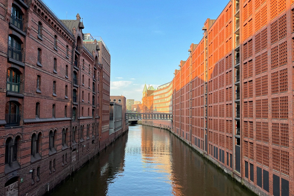 Schmaler Kanal in der Speicherstadt flankiert von historischen roten Backsteinlagerhäusern mit Giebeln links und modernerem Neubau rechts, Morgenlicht